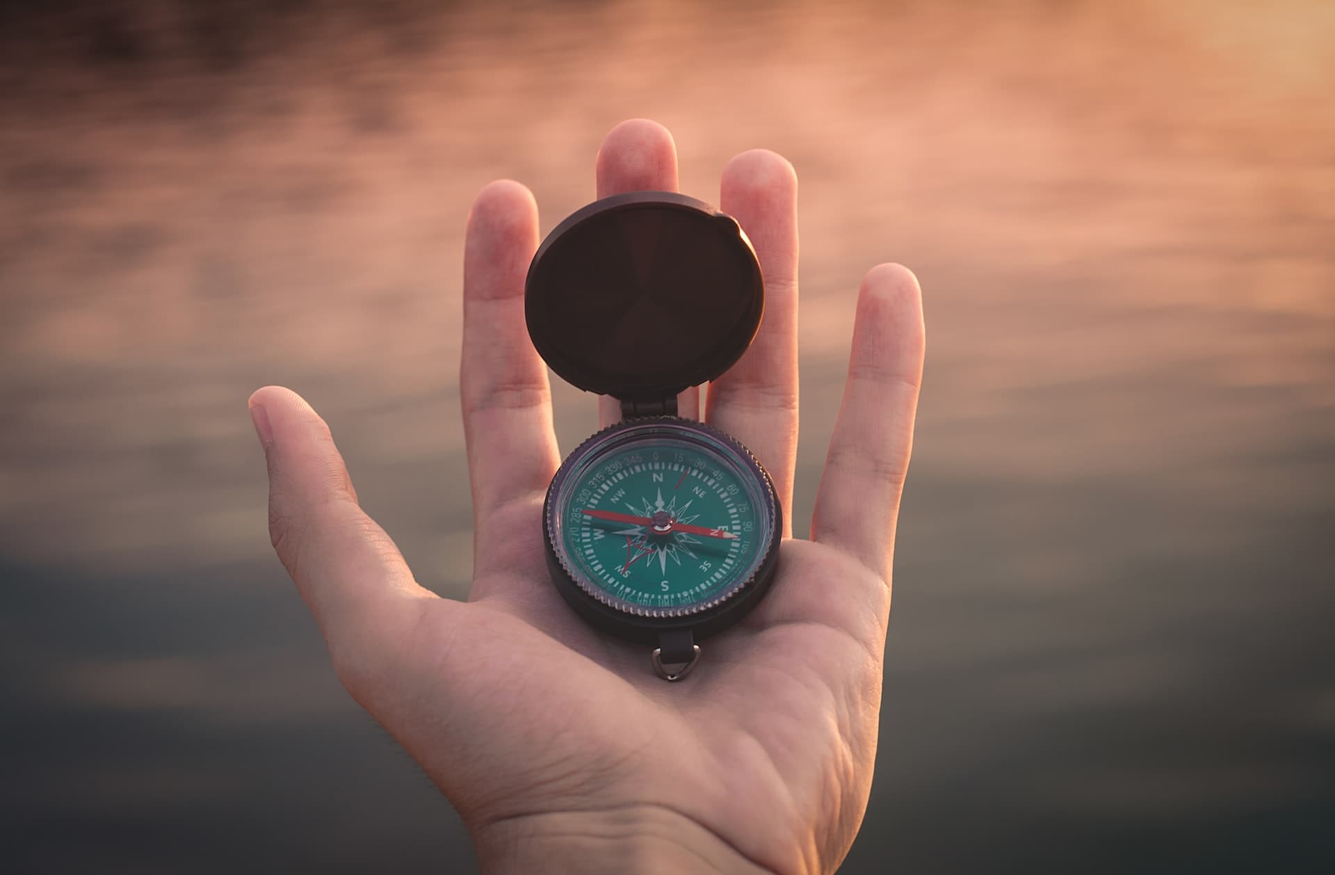 A hand holding a compass, sunset light reflecting on water in the background
